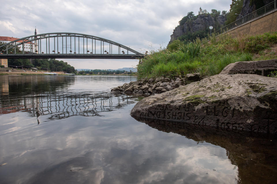 A large rock in the Elbe River, with a bridge in the background. is carved with a deeply incised inscription in what appears to be archaic lettering.