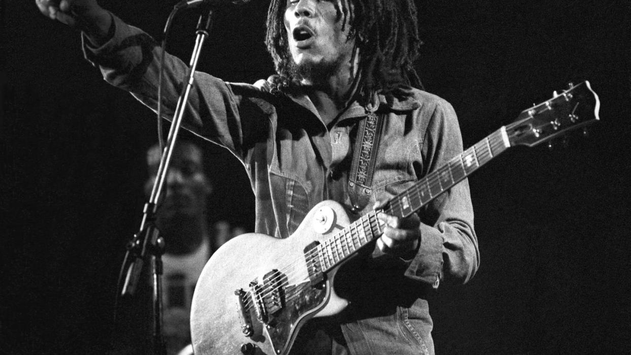 bob marley playing guitar and singing at a microphone and pointing offstage