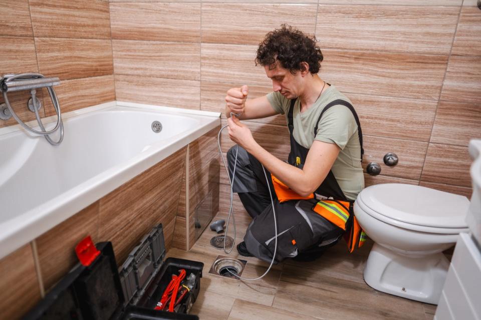 A man in overalls uses a tool to fix a drain in a bathroom.