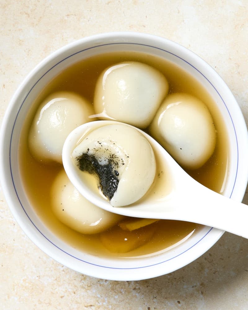 Bowl of tang yuan dessert rice balls stuffed with sesame paste in a sweet broth with one on a spoon with a bite taken out.