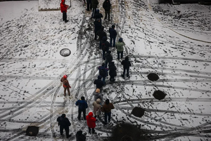 More than a dozen people wait in line in the snow outside a supermarket.