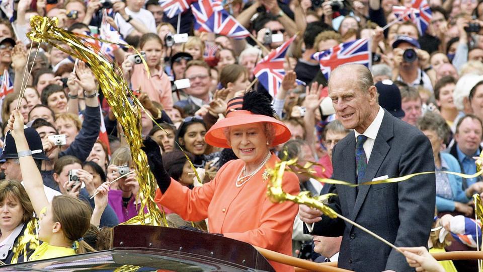 queen elizabeth ii and prince philip stand in the bed of a car that travels through crowds, both smile and wave as people wave british flags and golden streamers, the queen wears an orange outfit and matching hat, the prince wears a gray suit