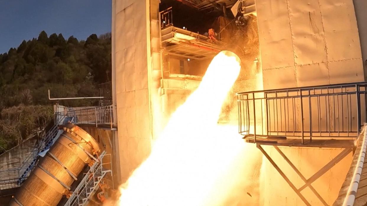  Flames shoot out of a cone-shaped rocket engine poking out of a hangar-like building. 