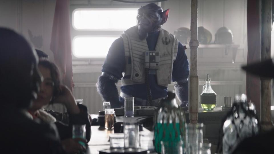 The large, big eared purple Lasat Zeb in his flight suit stands at a bar on The Mandalorian