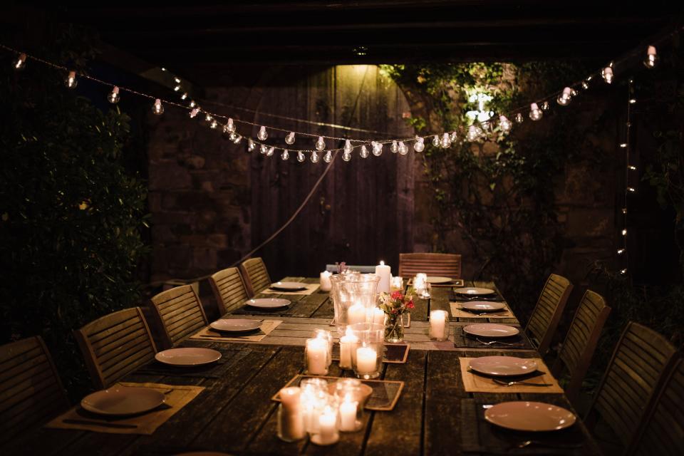 A long dinner table with set places and candles. String lights with a soft, warm hue are hung overhead.