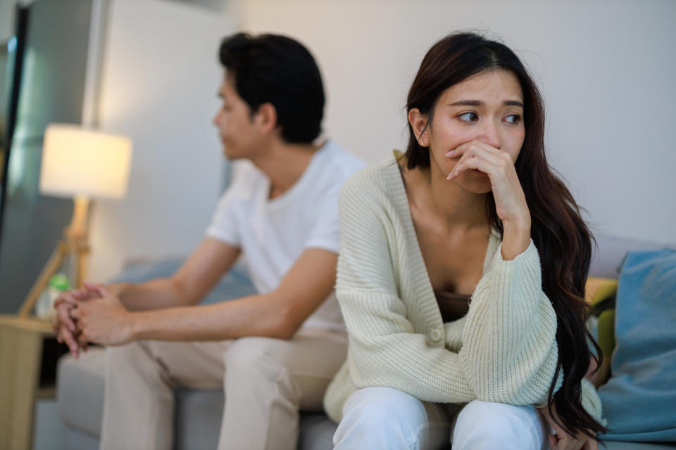 A man and woman sit on a couch appearing upset, facing away from each other. The woman has her hand on her mouth, while the man looks down with his hands clasped