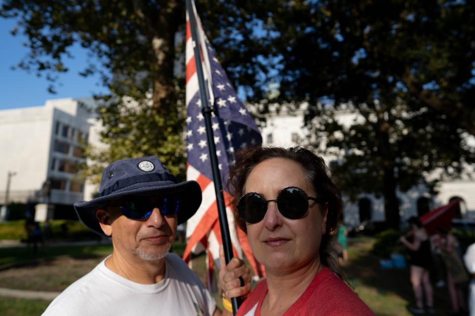 Two people face the camera with an American flag behind them.