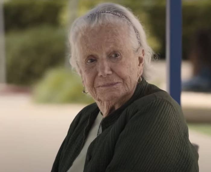 Elderly woman sitting outdoors, smiling gently, wearing a scarf and cardigan