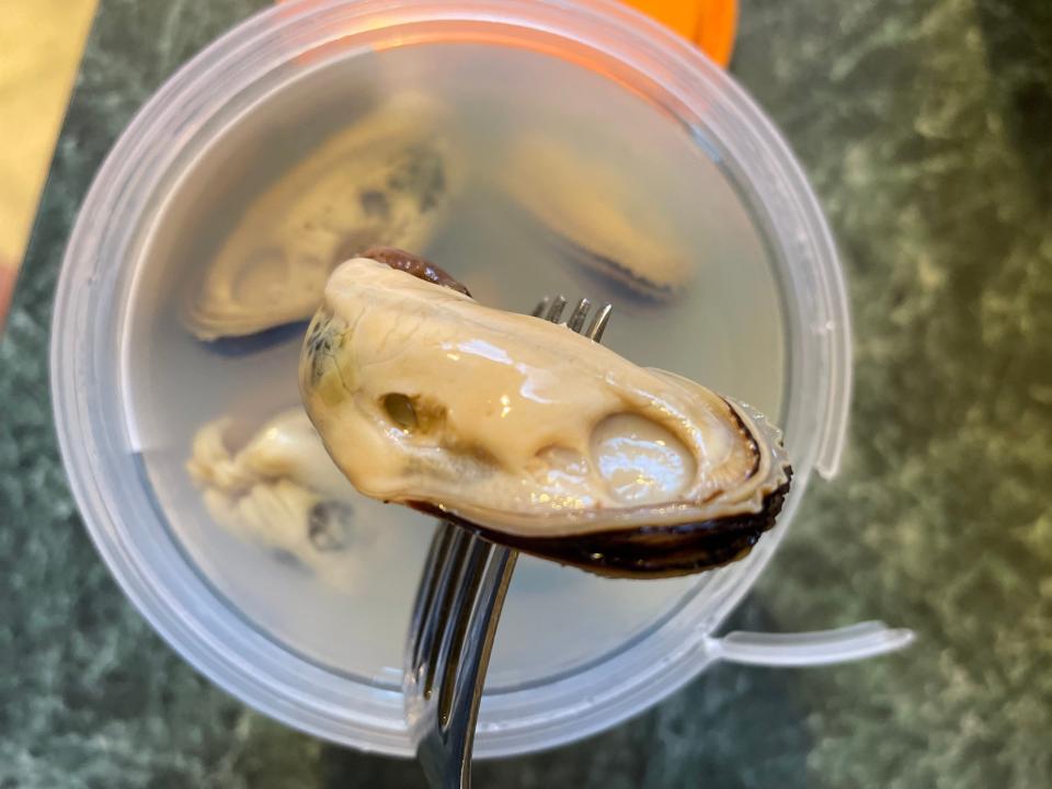 A fork stabbed through a marinated mussel with a container of mussels in a clear solution in the background