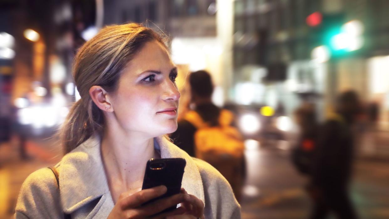  white woman with blonde hair pictured walking down a city sidewalk at night with her phone illuminating her face. She's looking up at the surrounding scene as if confused 