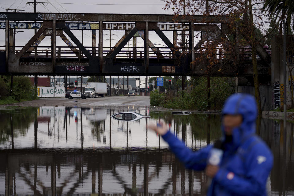 A television reporter does a broadcast as cars are seen submerged on a flooded street under a railroad bridge Thursday, Feb. 1, 2024 in Long Beach, Calif. (AP Photo/Eric Thayer)