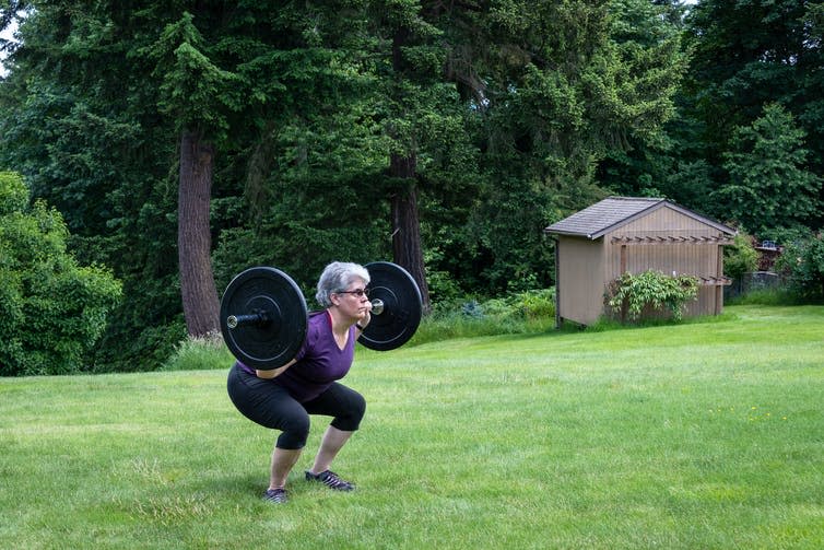 An older woman performing a barbell squat outdoors.