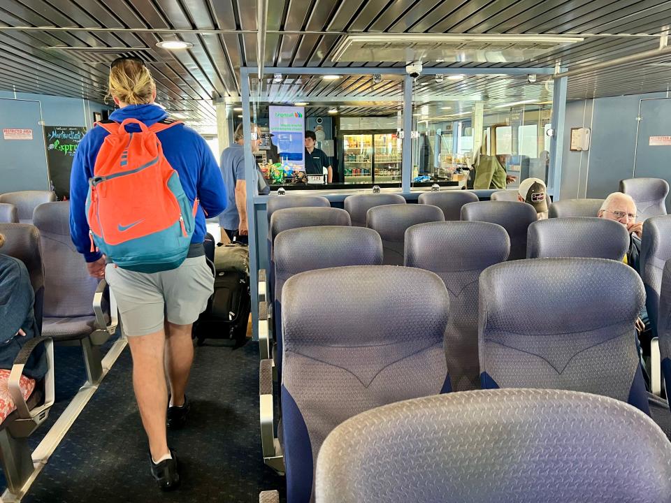 Several black cushioned seats on a ferry. A woman wearing a blue hoodie and orange and blue backpack walks along the aisle.