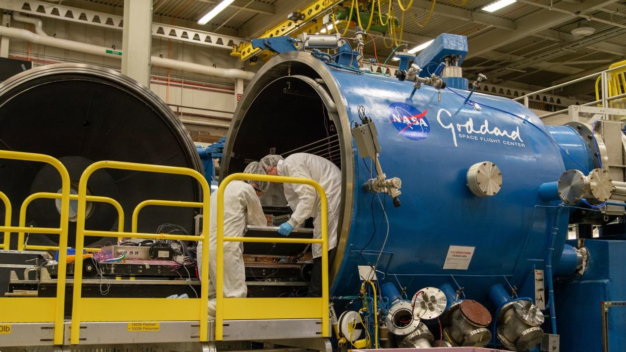  Scientists bring the ComPair instrument into a blue vacuum chamber for thermal testing. The Goddard logo is on the side of the chamber and the scientists are wearing white sterile uniforms. 