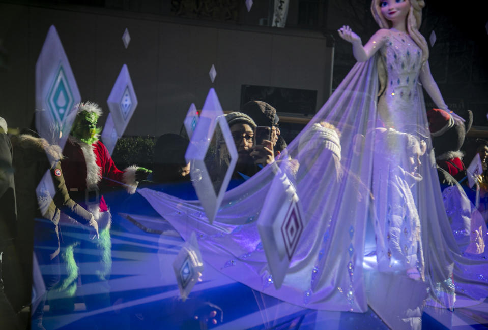 In this Friday, Dec. 20, 2019 photo, a costumed Grinch performer, left, works a sidewalk for tips near a department store holiday window display near Rockefeller Center in New York. Some performers, who solicit tips from tourists from designated "activity zones" in Times Square, have been migrating to Rockefeller Center for the holiday season where no such zones exist. (AP Photo/Bebeto Matthews)