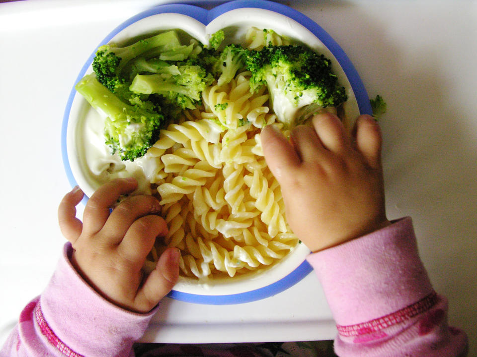 A child's hands playing with a plate of pasta and broccoli