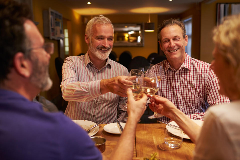 Four men smile and toast at a dinner table in a restaurant. One of the men has a beard and another has curly hair. Their names are not provided