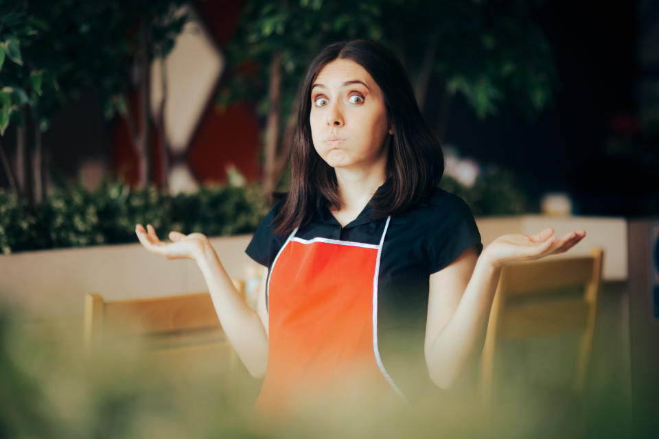 A woman wearing a red apron and black shirt stands in a restaurant, shrugging with a confused facial expression