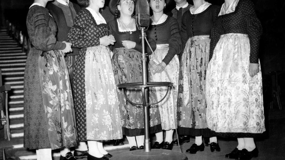 eight members of the von trapp family stand around a large microphone and sing, the girls wear dresses with matching aprons and hats, the men wear matching tops