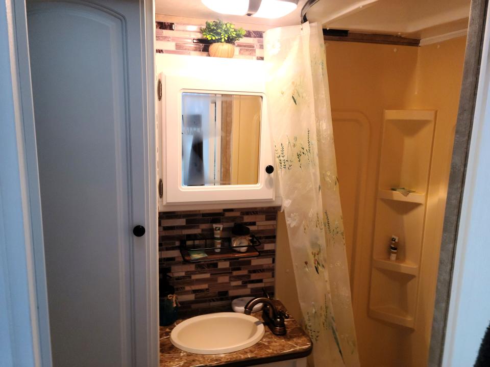 A small bathroom with a sink, medicine cabinet with a mirror, overhead light, shower with three shelves, and a floral shower curtain.