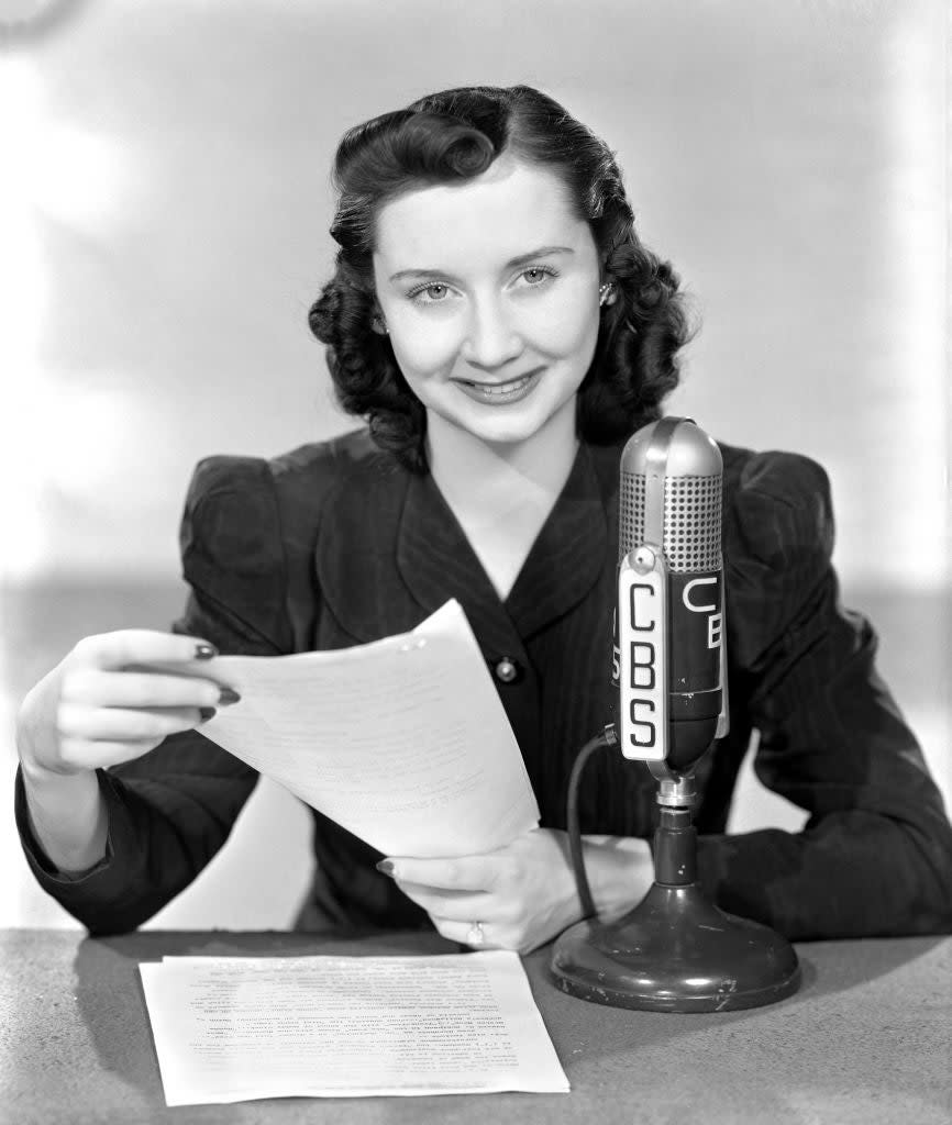 Woman holding papers and smiling while sitting at a desk with a CBS microphone