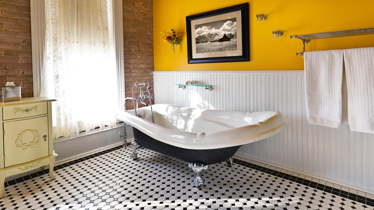 A modern contemporary classic bathroom design, furnished with a classic painted cabinet, sepia toned picture on the wall, exposed brick wall and a window, a claw foot bath tub and black and white tile pattern on the floor.