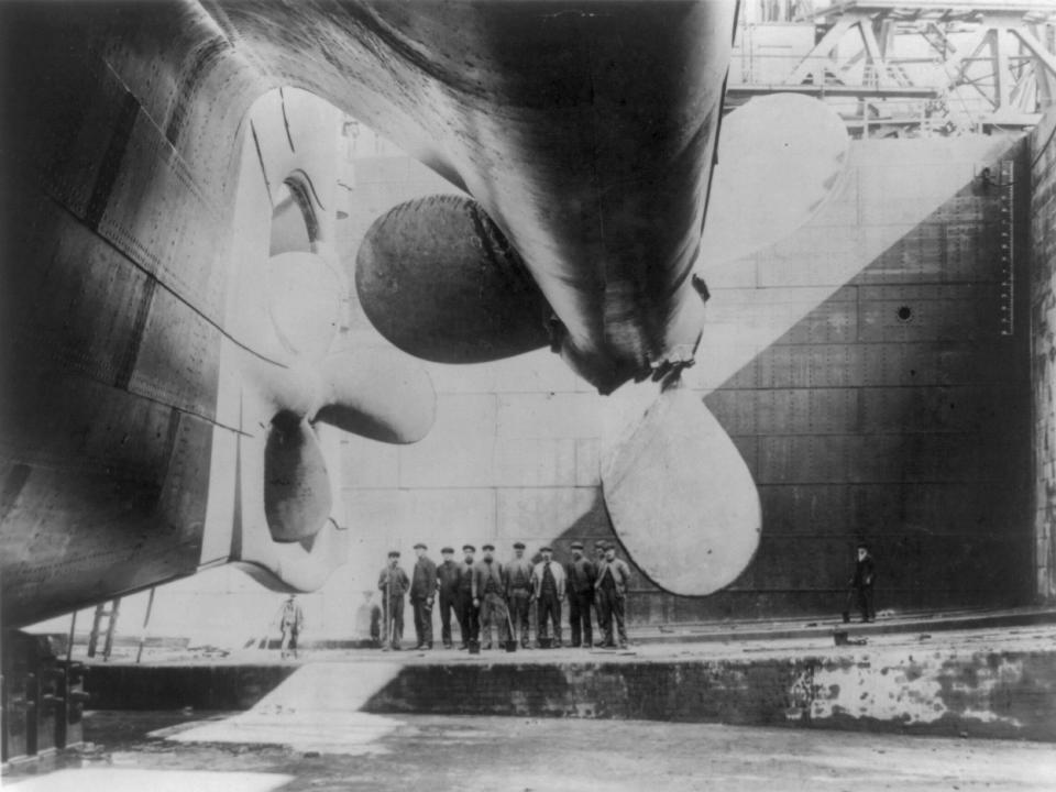 A black-and-white image of a group of people standing and looking at the propellor at the back of the Titanic.