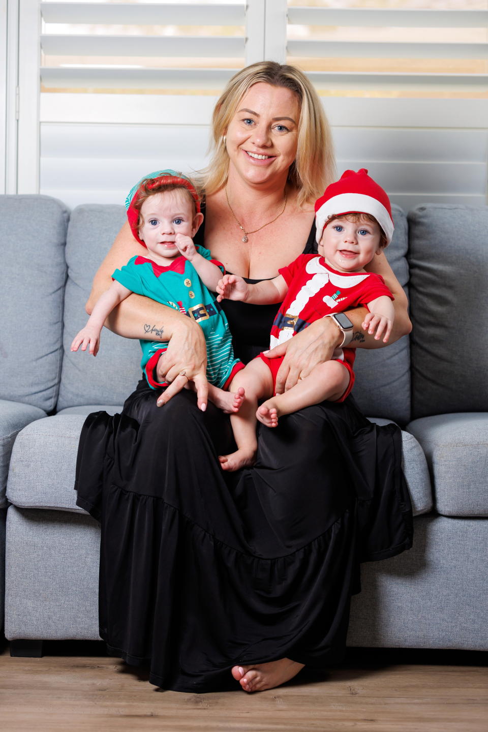 Casey Edwards holding twins Lucas (left) and Bailey (right) dressed as Santa and an elf on a couch at their home. 