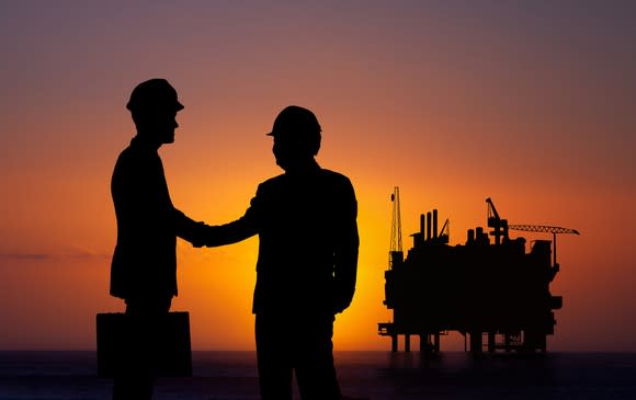 The silhouette of two people shaking hands with an offshore oil platform in the background.