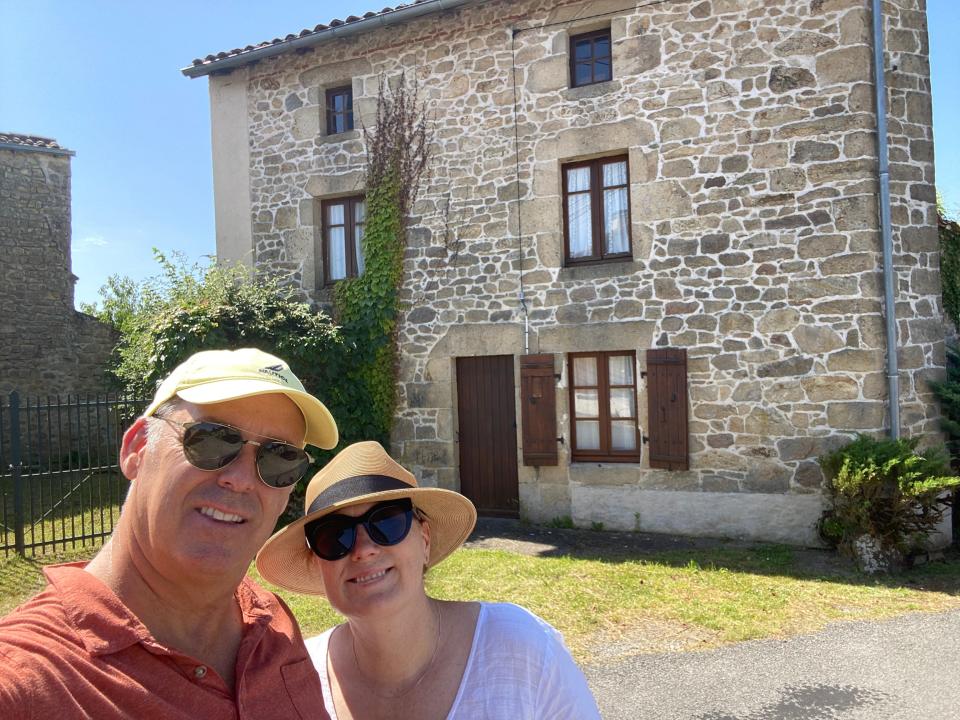 A Gen X couple posing in front of their stone cottage in the French countryside.