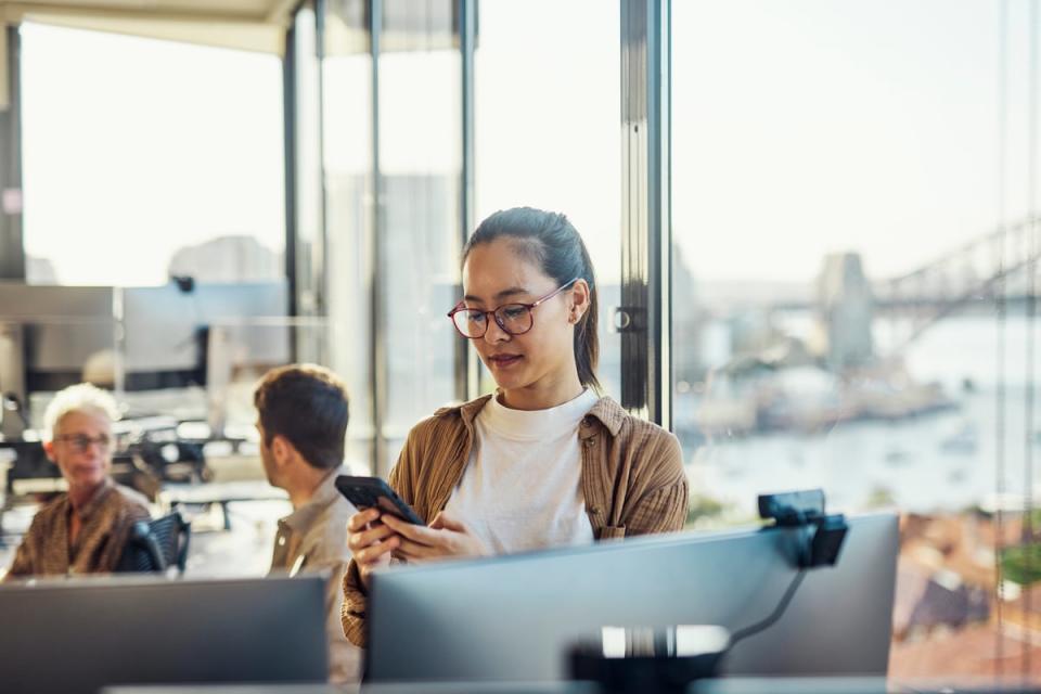 A person standing in an office setting looks at their cell phone in front of two computer monitors. 