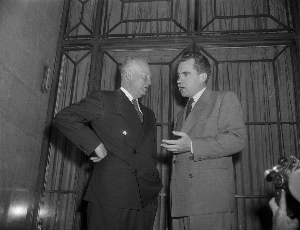 Two men in suits gesture toward each other in a black and white photo.