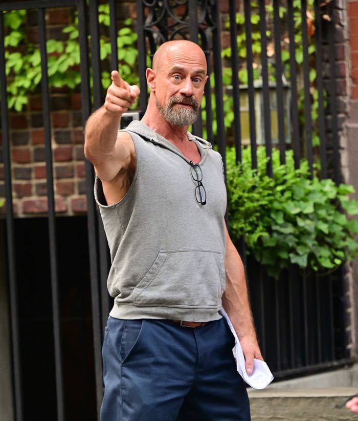 Chris Meloni outside in a cutoff sweatshirt and pants pointing at the camera, script in hand