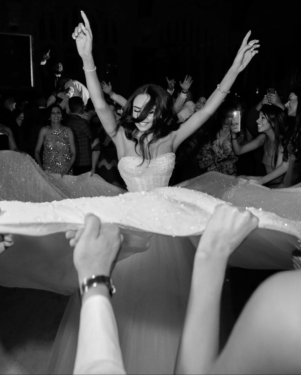 A black and white photo of a bride twirling as her guests hold up her wedding skirt.