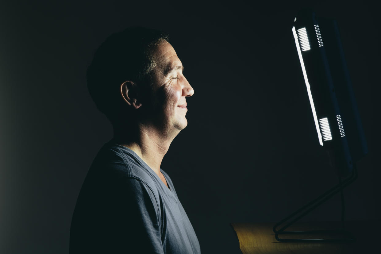 older man smiling standing in front of light therapy lamp in a dark room, seasonal affective disorder light therapy lamp