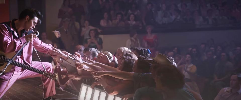 Elvis kneeling on stage holding hands of young women reach out to him