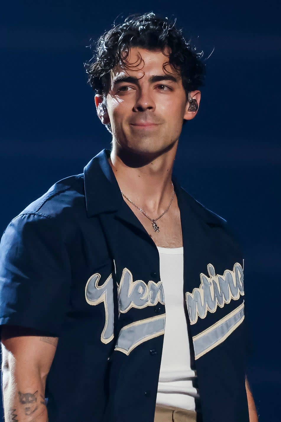 Joe Jonas in a varsity jacket and beige pants performing on stage with a microphone