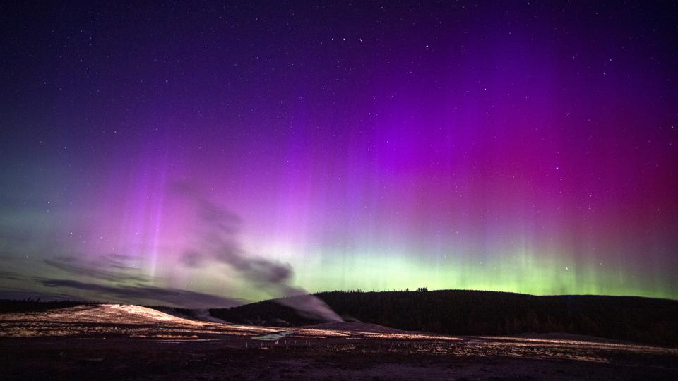  Northern lights appear as ribbons of purple and pink light with some green lower down on the horizon across the sky. stream from a geyser rises up in the foreground. . 