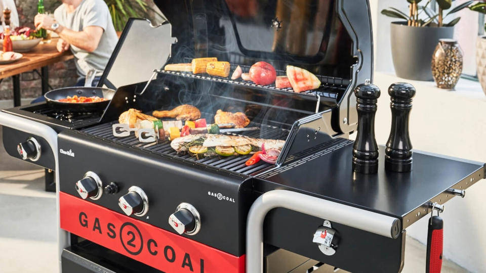 Gas grill cooking a selection of meats and vegetables, people sat around table in background