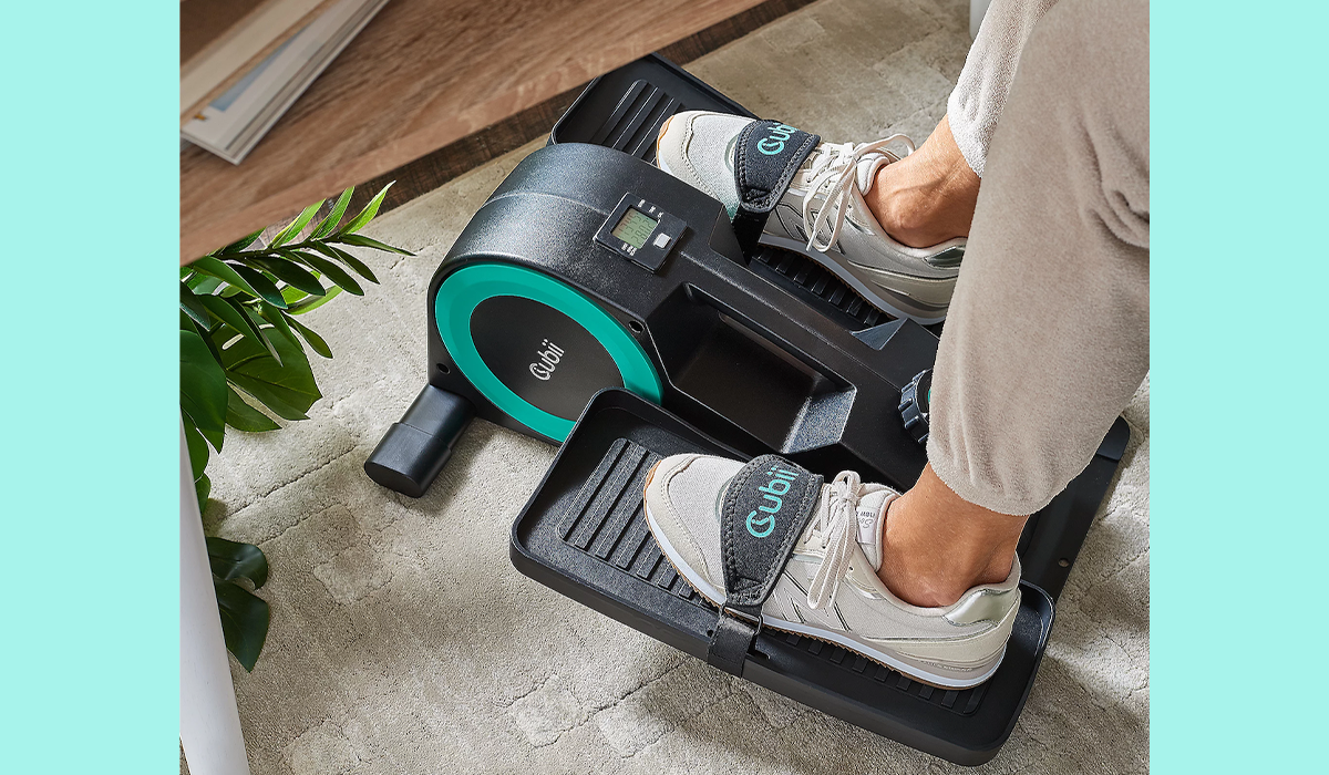 Photo of woman in sweats and sneakers with feet on Cubii machine below her desk.