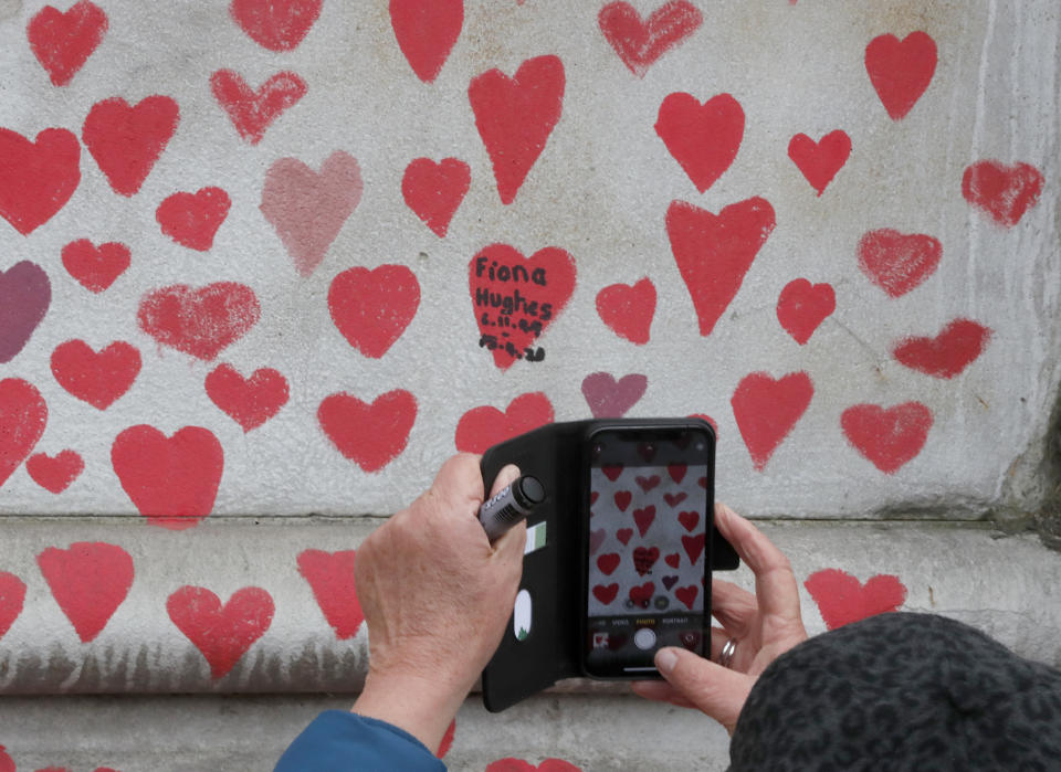 A woman takes a picture on her phone as people paint red hearts marking the completion of the approximately 150,000 hearts being painted onto the National Covid Memorial Wall to commemorate all those who have died of coronavirus, on the Thames Embankment opposite the Houses of Parliament in London, Thursday, April 8, 2021. Bereaved families want the wall of painted hearts to remain a site of national commemoration and are asking the Prime Minister to help make the memorial permanent. (AP Photo/Frank Augstein)