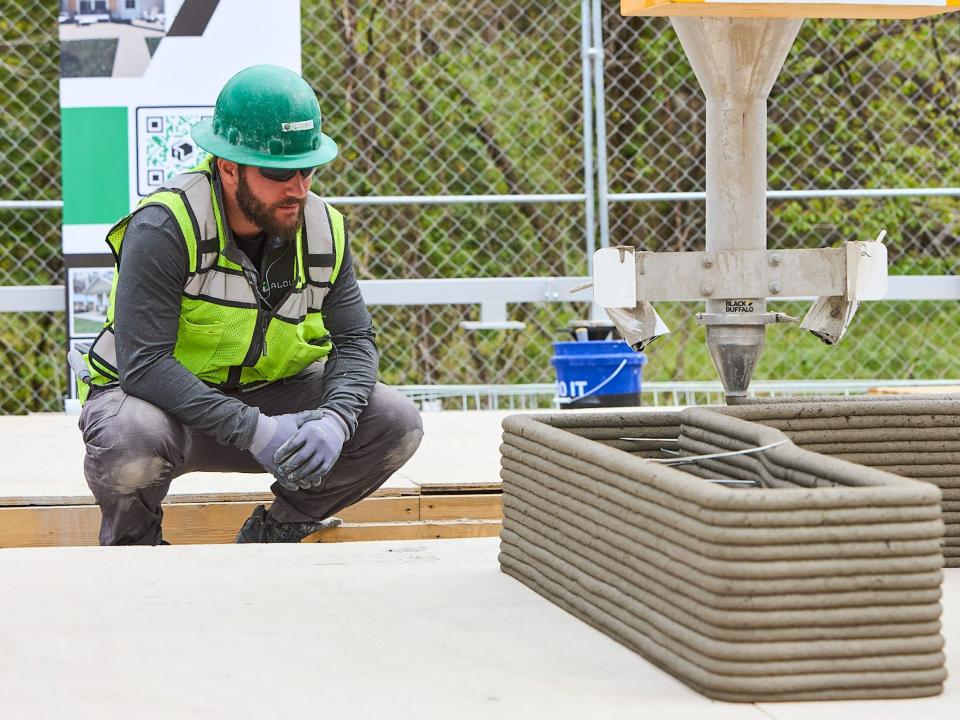 A 3D printer printing concrete onto a wall. A person is kneeling next to the printer.