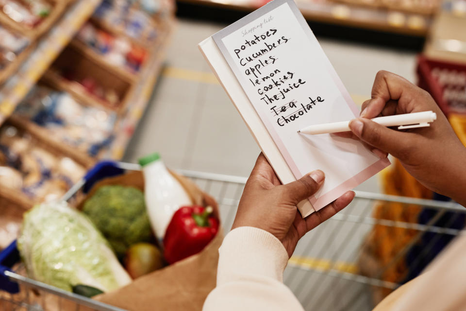 A person holding a shopping list with items like potatoes, cucumbers, lemons, cookies, tea, and chocolate, while pushing a shopping cart with groceries