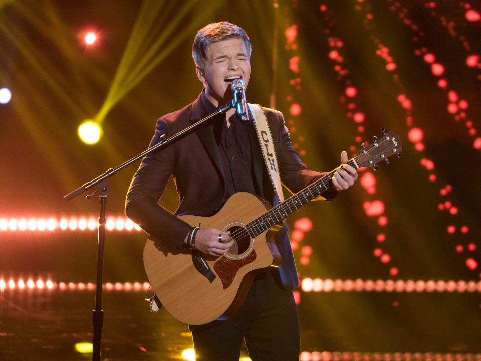 Caleb Lee Hutchinson in a brown suit playing a guitar and singing onstage with red lights and illuminated background