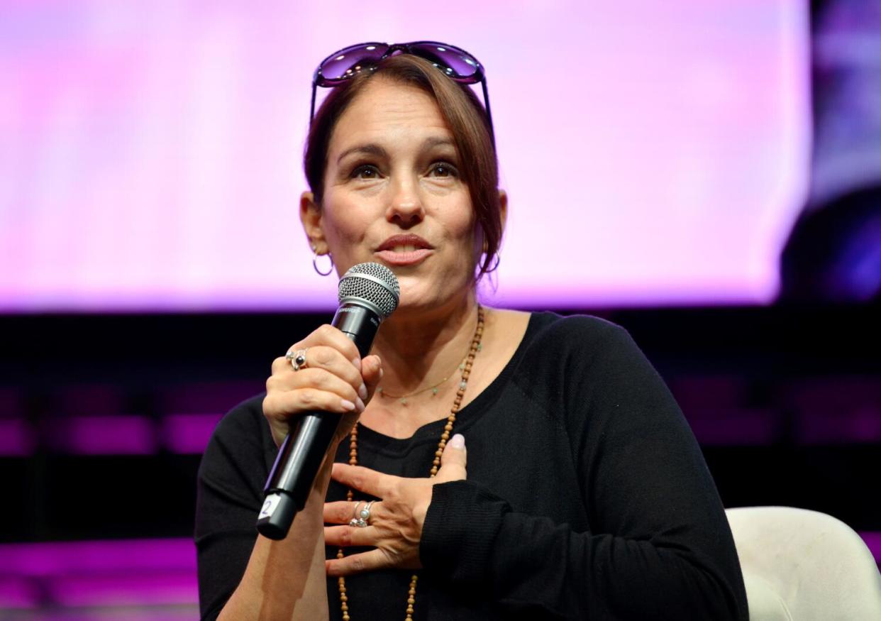 A woman in a black top gestures while speaking at a microphone.