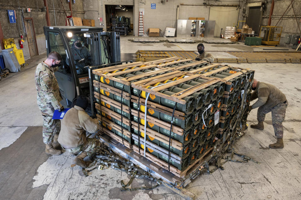 Several people in military fatigues attend to weaponry stacked on a forklift in what appears to be a mostly empty warehouse.