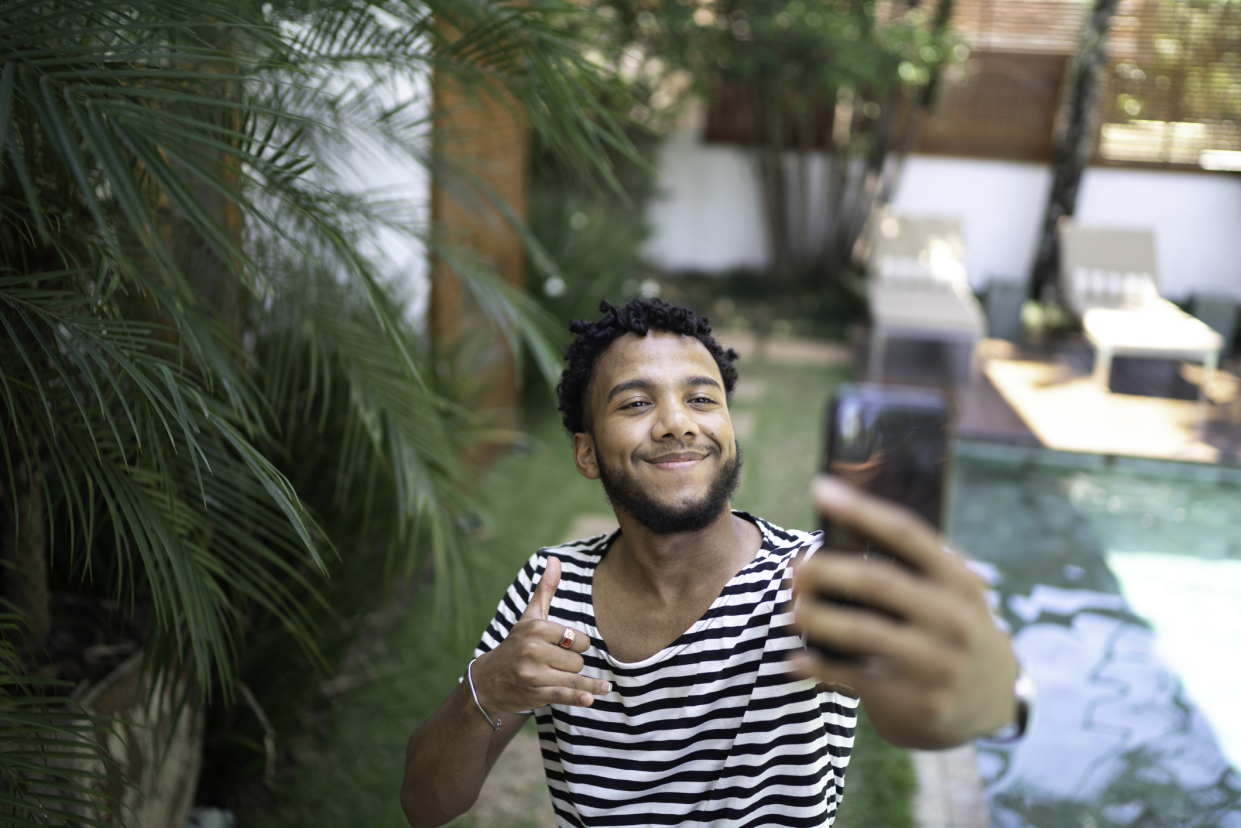 Young man smiling holding his smartphone with left arm stretched, taking a video of himself, outside in his yard, patio and swimming pool on the right, a palm tree in the foreground on the left