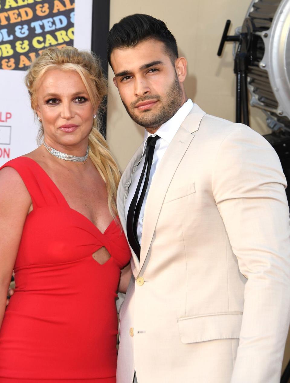 britney spears and sam asghari, a young couple hugging and looking at the camera, the woman wears a red dress with blonde hair in a ponytail, the man wears a cream suit with black hair slicked back