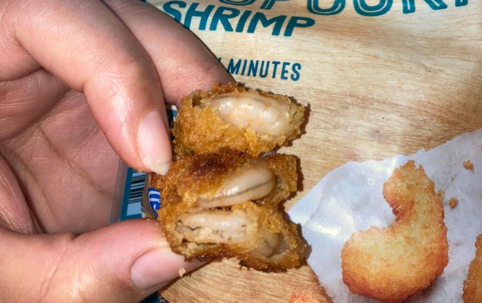 Woman's hand holds battered prawns with a thin, black string down their back in front of packet of Aldi Fremont Fish Market Oven Crispy Popcorn Shrimp