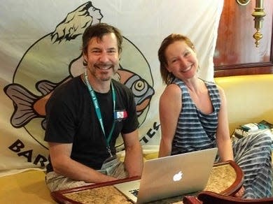 Kat Whitmire and her husband sitting and smiling in front of a tapestry of a person riding a fish — the logo of the cruise company Bare Necessities.